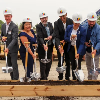 Six people in business attire and hard hats hold shovels, posing for a groundbreaking ceremony at a construction site.