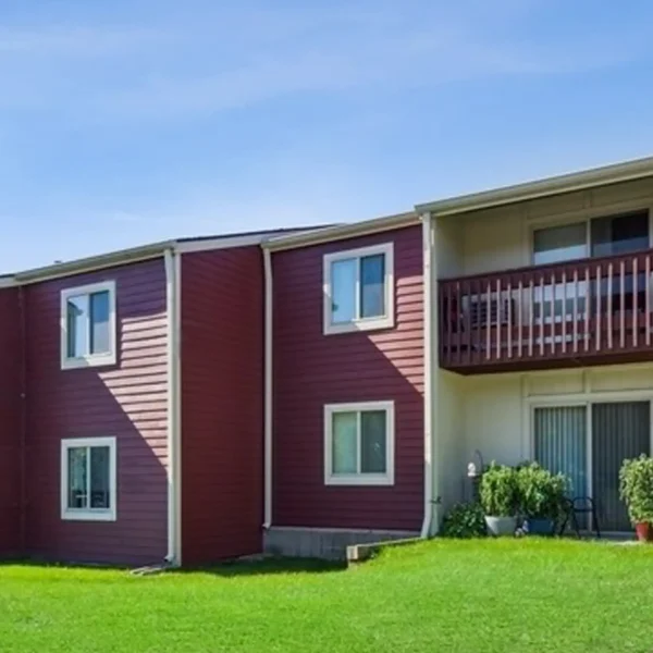 Two-story apartment building with red siding, white trim, balconies, and windows, situated on a grassy lawn under a clear blue sky.