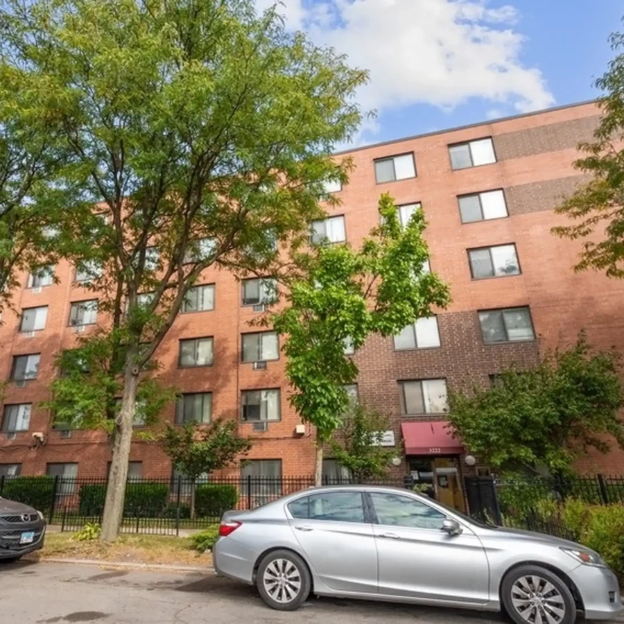 A four-story red brick apartment building with several trees and parked cars in front, photographed on a partly cloudy day.