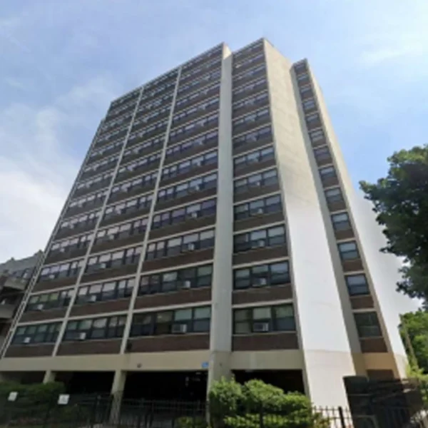 A multi-story residential apartment building with numerous windows, brown and white exterior walls, and a small fenced area in front.