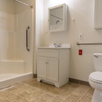 Accessible bathroom with a walk-in shower, grab bar, white vanity with mirror, toilet, and beige tiled floor.
