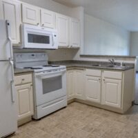 A kitchen with white cabinets, a refrigerator, stove, microwave, sink, and gray countertop on tan tile flooring.