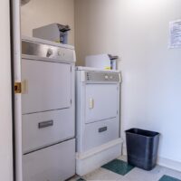 Two white coin-operated laundry machines, a washer and dryer, are placed side by side in a small room with white walls and a black trash bin nearby.