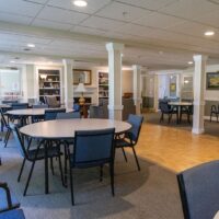 A spacious, well-lit room with round tables and blue chairs, bookshelves, and artwork on the walls, designed for group activities or meetings.