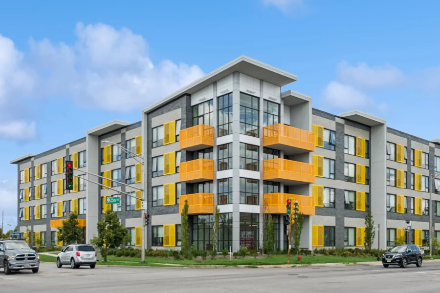 A modern four-story apartment building with gray and white exterior accents, yellow balconies, and large windows at a street intersection with cars and traffic lights.
