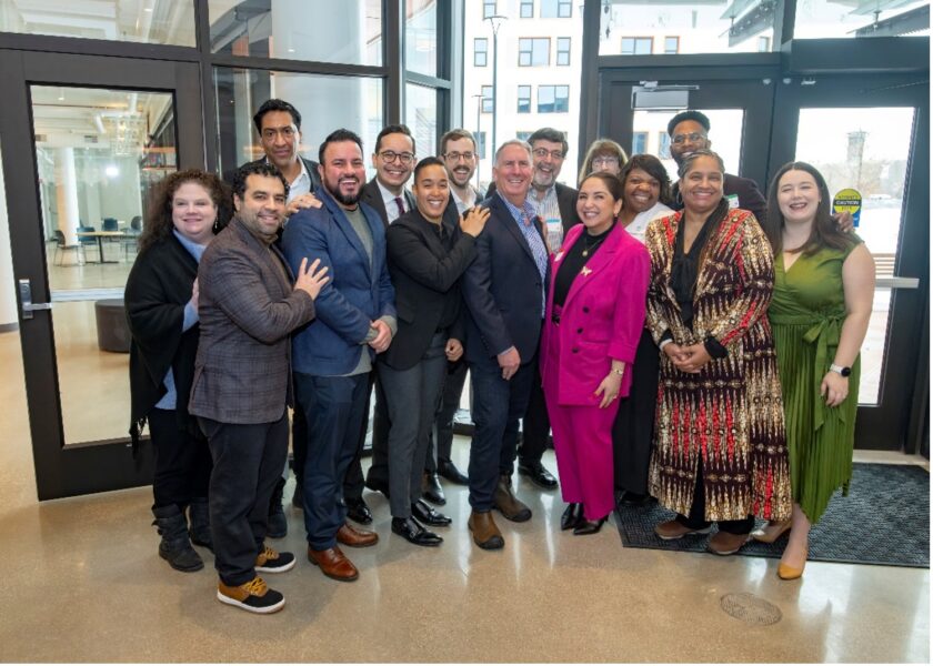 A group of fifteen people stand together indoors, dressed in business and semi-formal attire, smiling and posing for a group photo near large glass doors.