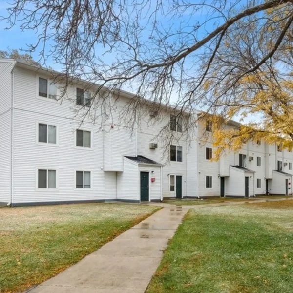 A three-story white apartment building with multiple entrances, surrounded by a grass lawn and trees, under a partly cloudy sky.