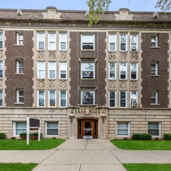 A four-story brown brick apartment building with decorative stone details, symmetrical windows, a central arched entrance, and a small lawn in front.