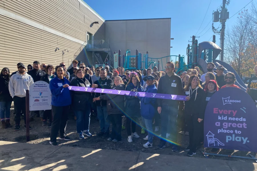A group of people stands together at a ribbon-cutting ceremony in front of a playground, with a sign that reads, "Every kid needs a great place to play.