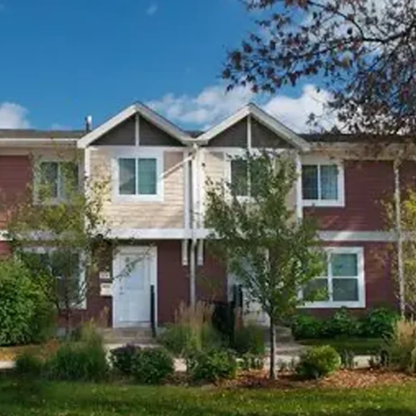 A two-story townhouse with red and beige siding, white trim, and a landscaped front yard with trees and shrubs.
