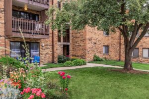 A brick apartment building with balconies, a green lawn, a tree, sidewalks, and a garden bed with colorful flowers in front.
