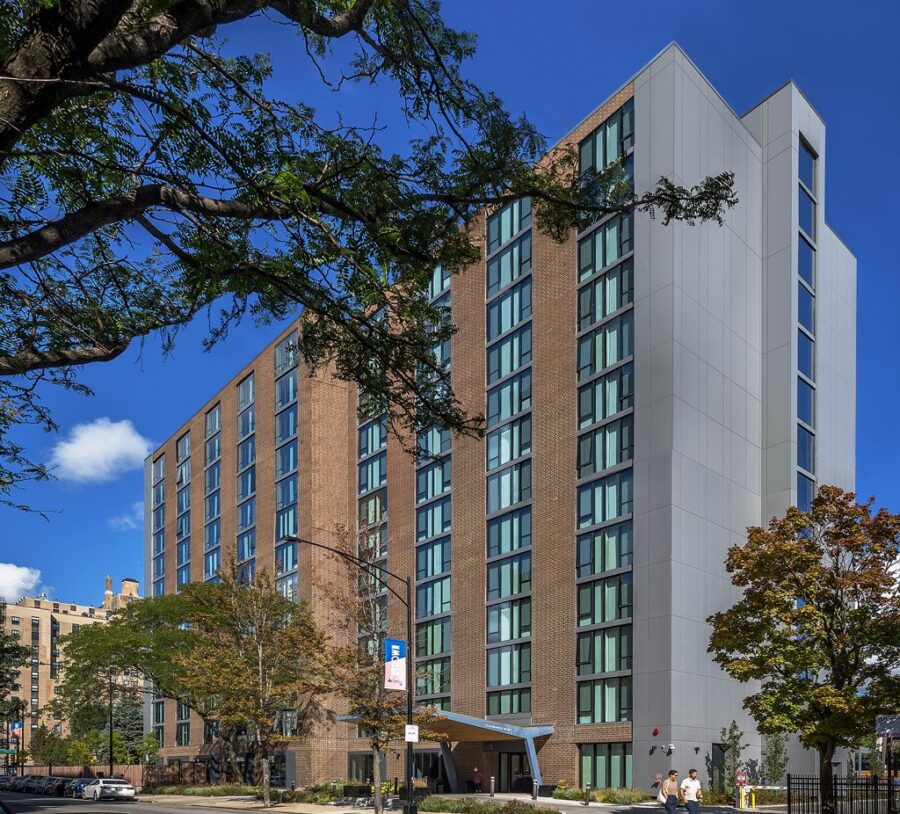 A mid-rise modern hotel with large windows stands on a city street corner, surrounded by trees and pedestrians, under a clear blue sky.
