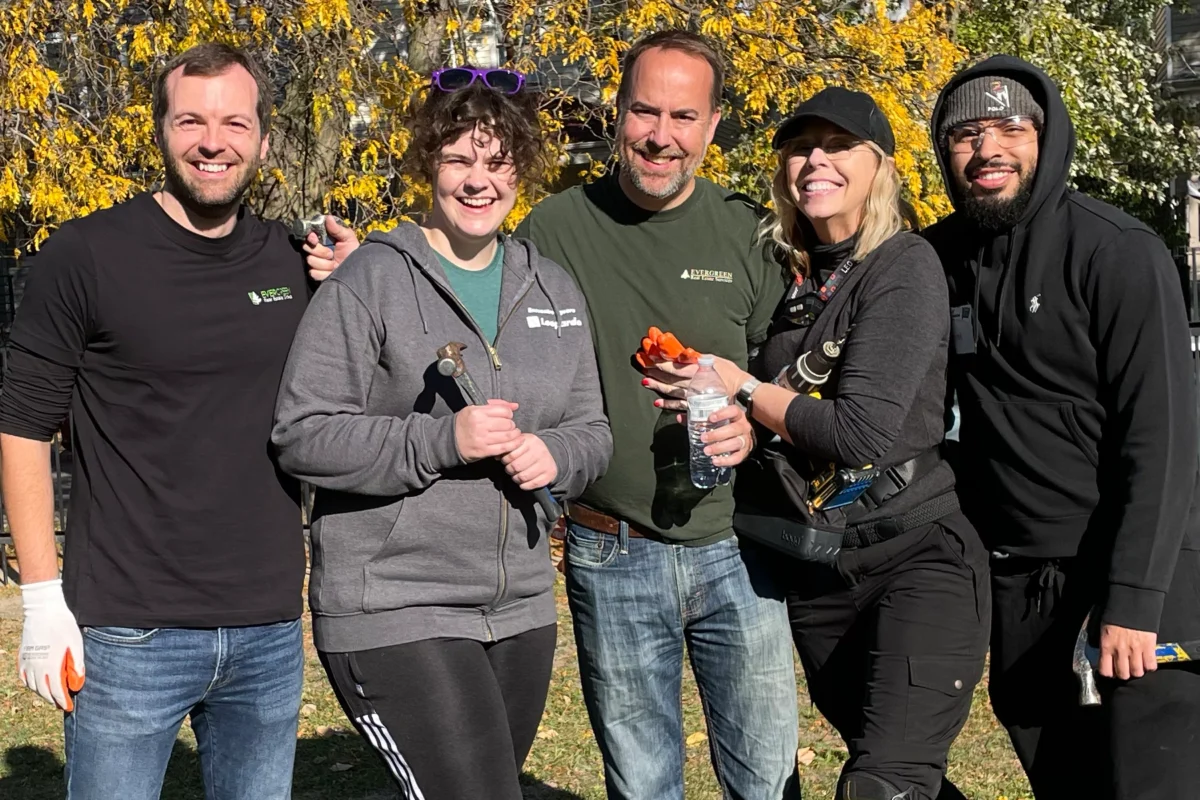Five adults stand outdoors in casual clothing, smiling at the camera, with yellow autumn foliage in the background.