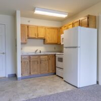 A small kitchen with wood cabinets, a white refrigerator, stove, microwave, and stainless steel sink. The floor is tile, and carpet is visible in the adjacent area.