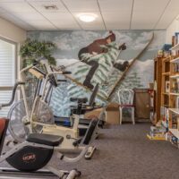 Exercise bikes and equipment are arranged in a room with a mural of a skier on the wall; bookshelves and board games are visible on the right side.