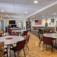 Community room with round tables and red chairs, a pool table, wall-mounted TV, refrigerator, and American flag on the wall.