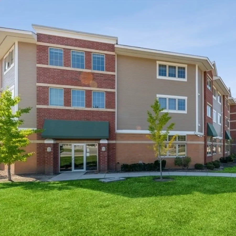 Three-story brick and siding building with green awnings, large windows, and a glass entrance door, surrounded by a grassy area and young trees.
