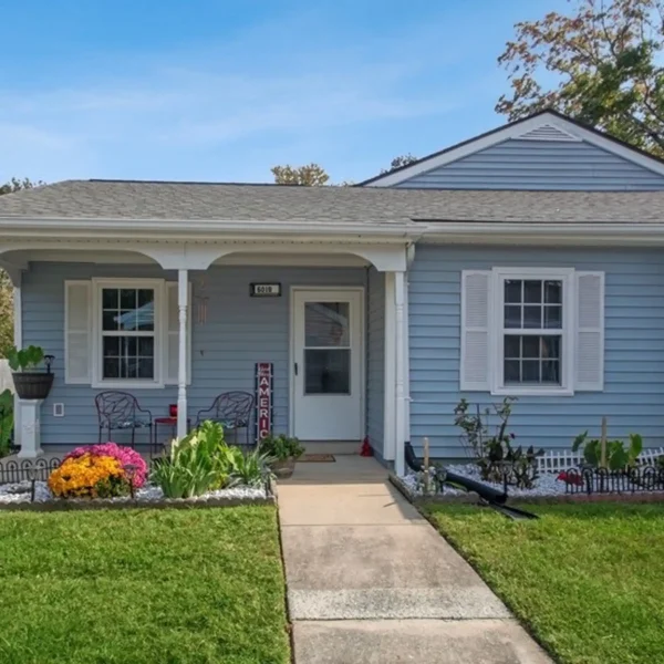 Single-story blue house with white trim, small covered porch, outdoor chairs, potted plants, and a walkway leading to the front door. Manicured lawn and flowers in the front yard.