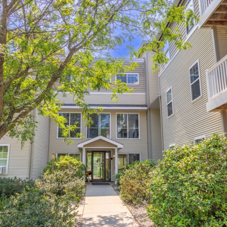 Entrance to a modern, three-story apartment building with beige siding, large windows, and a pathway leading through landscaped greenery.