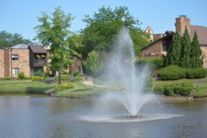 A water fountain sprays in the center of a pond, surrounded by grass, trees, and brick buildings under a clear blue sky.