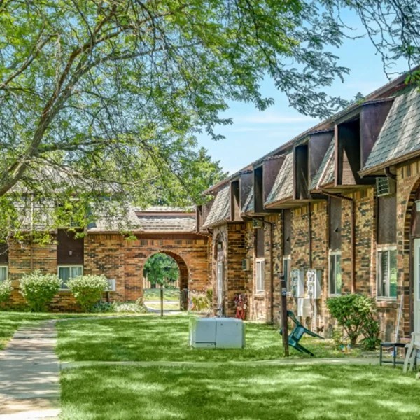 A brick apartment building with brown roof accents is shown along a green lawn and walkway, with trees providing shade and an arched entryway visible in the background.
