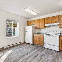 A kitchen with wood cabinets, white refrigerator, white stove with range hood, and a window showing a playground outside. The floor is gray wood-look vinyl.