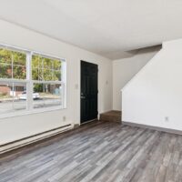 A room with wood-look vinyl flooring, white walls, a large window, a black door, and a staircase leading upward.