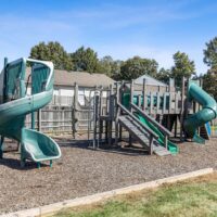 Outdoor playground with green slides, climbing structures, and wood chip ground cover, surrounded by a wooden fence and trees in the background.