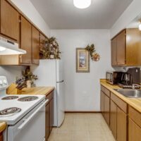 A small kitchen with wood cabinets, white appliances, a countertop with kitchen gadgets, and decorative plants on top of the refrigerator and cabinets.