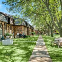 A shaded sidewalk runs between brick apartment buildings with grass lawns, large trees, outdoor furniture, and various utility units on a sunny day.