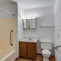 A small bathroom with a bathtub and grab bar, a vanity with sink, mirrored medicine cabinet, toilet, and wall shelf, all with white walls and wood flooring.