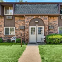 A brick apartment building with two white doors under an arched entryway, flanked by chairs and greenery, and a concrete path leading up to the entrance.