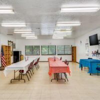 A community room with tables covered in red, white, and blue tablecloths, chairs, an American flag, bookshelves, large windows, and a wall-mounted TV.