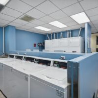 A laundromat room with blue walls contains several top-loading washing machines in the foreground and stacked dryers in the background under fluorescent lighting.