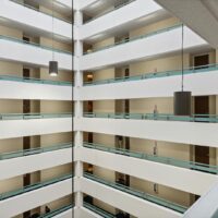 Interior view of a multi-story building with open hallways, turquoise railings, beige walls, and hanging cylindrical pendant lights.