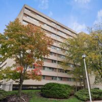 A mid-rise beige apartment building with multiple windows, surrounded by trees with autumn foliage and trimmed bushes, under a partly cloudy sky.
