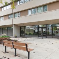 A wooden bench sits on a concrete walkway outside a building with large glass windows and doors; scattered leaves are on the ground.