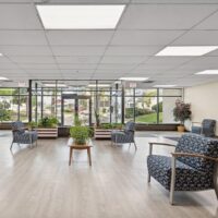 A bright waiting area with patterned armchairs, potted plants, large windows, light wood flooring, and views of greenery outside.