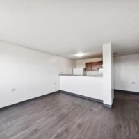 A small, empty apartment room with white walls, gray wood flooring, and a partial view of a kitchen with brown cabinets and white appliances.