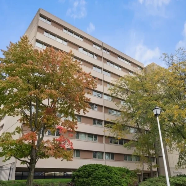 A mid-rise beige apartment building with multiple balconies, surrounded by trees and bushes, under a blue sky.