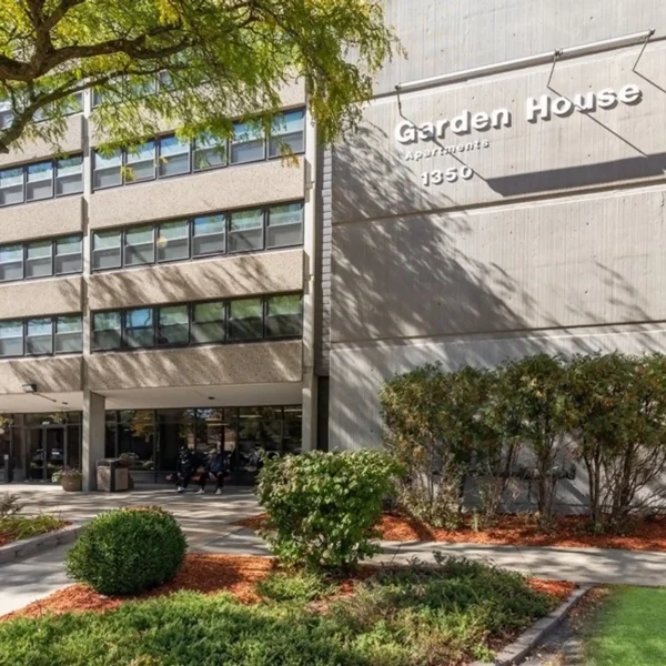 The entrance of a multi-story apartment building labeled "Garden House Apartments 1350," with landscaping and trees in front.