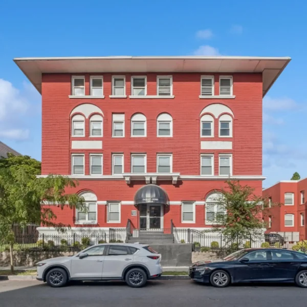 A four-story brick apartment building with arched windows and a central entrance canopy, seen from the street with two parked cars in front.