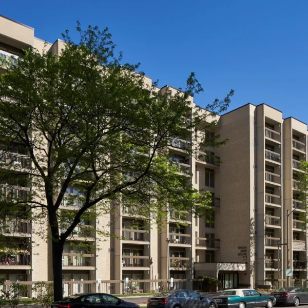 A multi-story apartment building with balconies, light brick exterior, and a large tree in front; several cars are parked along the street under a clear blue sky.