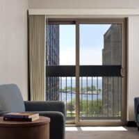 A sliding glass door with vertical blinds opens to a balcony overlooking buildings, trees, and a waterfront; an armchair and table with books are in the foreground.
