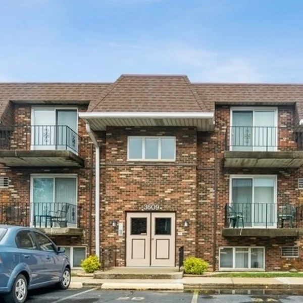 A two-story brick apartment building with balconies and a brown roof, featuring a central entrance and a parked blue car in front.