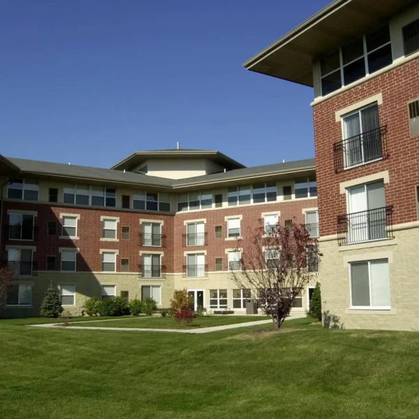 A modern, four-story apartment building with red brick and beige stone exterior, surrounded by a green lawn and small trees under a clear blue sky.