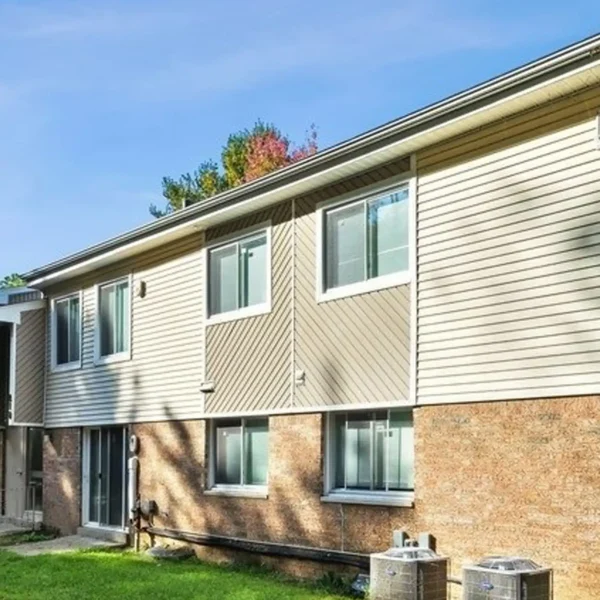 A two-story apartment building with beige siding and brick lower walls, featuring several windows and two outdoor air conditioning units on a grassy lawn.