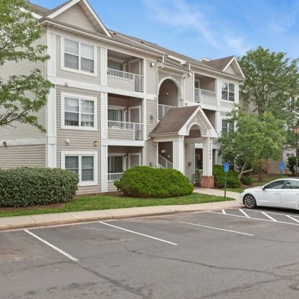Three-story apartment building with beige siding, white trim, balconies, and an arched entrance; a white car is parked in the lot with marked parking spaces and greenery around.
