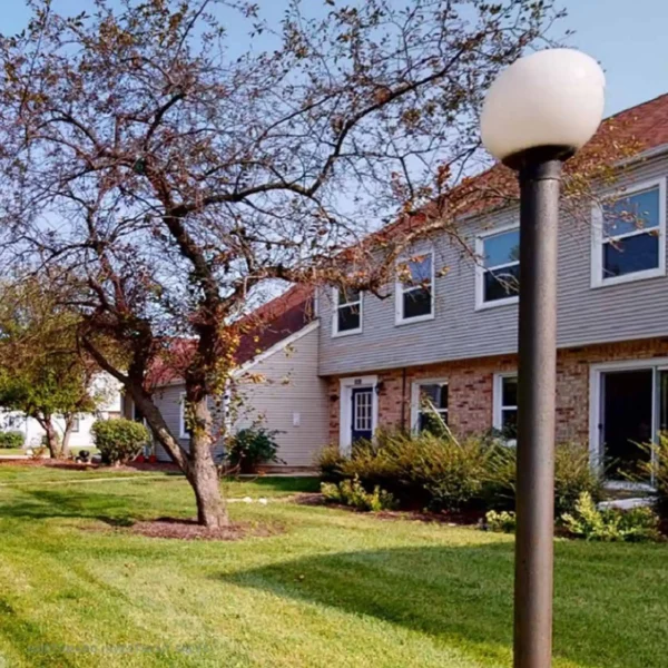 A grassy yard with trees, shrubs, and a round-topped lamppost in front of a two-story brick and siding residential building.