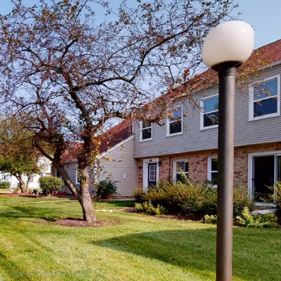 A grassy yard with trees, shrubs, and a round-topped lamppost in front of a two-story brick and siding residential building.